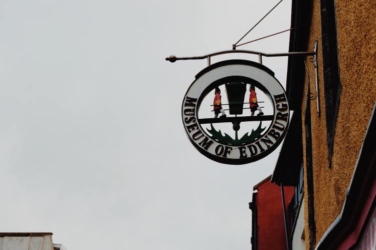 The circular sign post of the museum of Edinburgh against a grey sky. The sign has the name on the outer circle and in the centre has a thistle at the bottom and guards either side carrying a chest. The yellow of the museum of Edinburgh can be seen on the right of the photo.