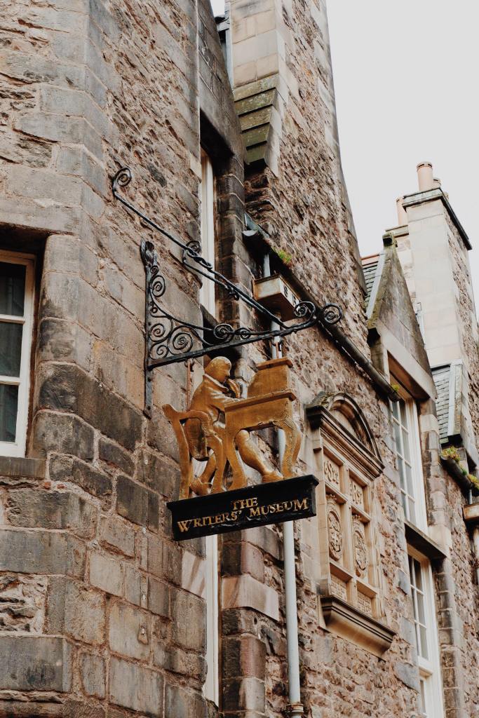 The metal sign post of the Writers museum, showing a man at a desk with a feather quill in gold and a black sign with The Writers Museum in gold lettering. Behind is the brown of the building, with windows. 