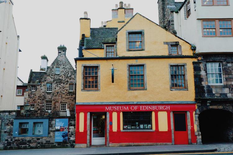 The exterior of the Museum of Edinburgh, from across the road. The top half of the building is two stories with yellow paint. The lower half is red with the name Museum of Edinburgh in white.