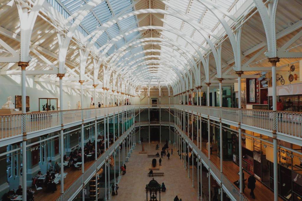 View over the main gallery of the National Museum of Scotland, from the top floor, looking over the three levels. The gallery has a glass roof and white posts around each of the balconies.