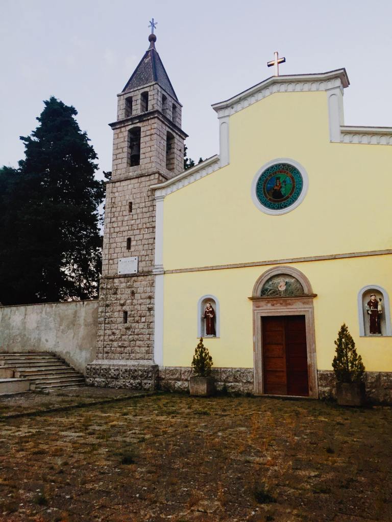 The convent of Santa Maria Delle Grazie in Jelsi, which is yellow with statues either side of a door and a circular stain glass window and cross on the top.