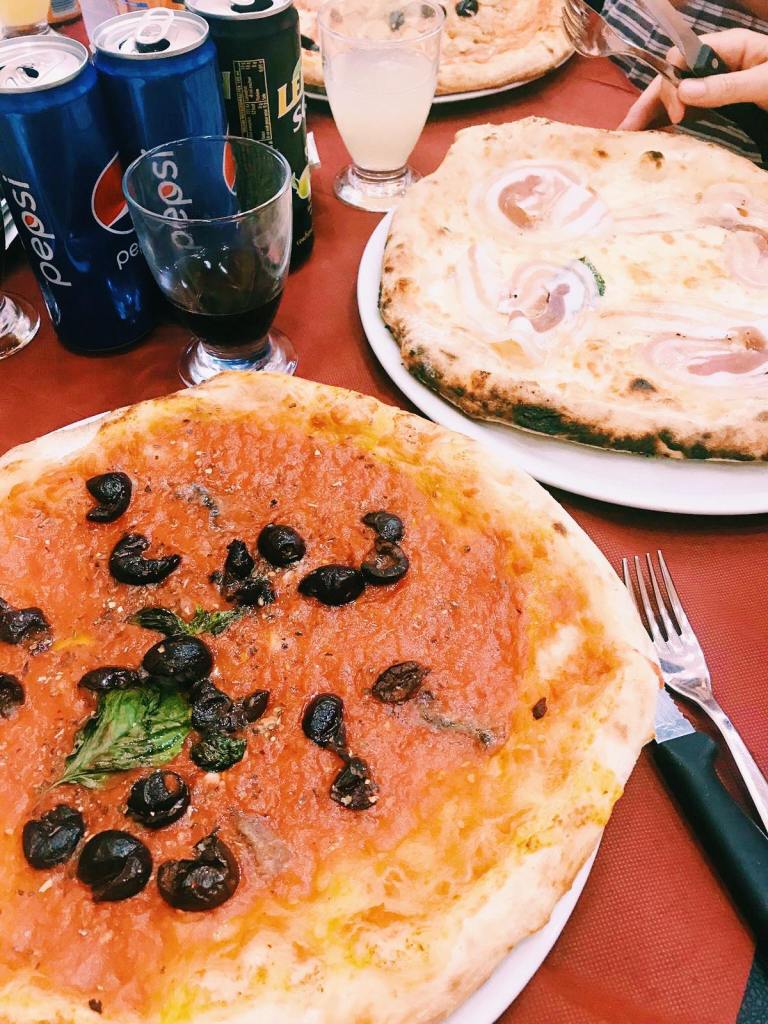 Marinara Pizza on a plate with black olives and basil, behind is a pizza with cotta and some Pepsi cans