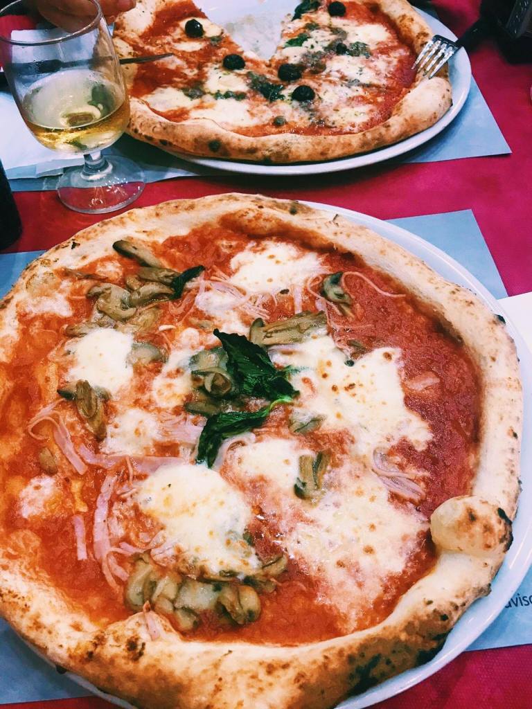 A large pizza with mozzarella, basil, shredded ham and mushrooms. Behind is a pizza with a slice cut out, with black olives and capers on it. A wine glass with white wine in between the Neapolitan pizza
