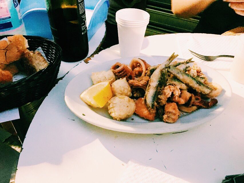 Plate of fried fish with lemon slice in the middle of a table with a bread basket nearby.