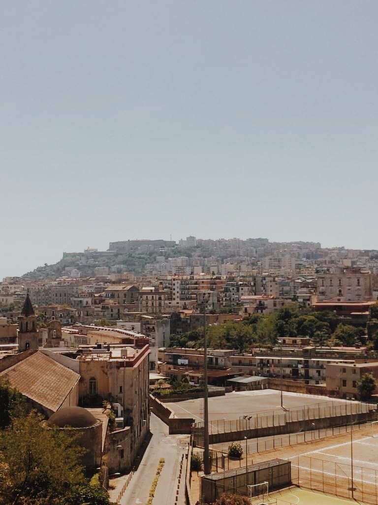 View of Naples, Italy from Cappodimonte, at the entrance to San Gennaro Catacombs. Image is filled with brown and yellow buildings up a hill.
