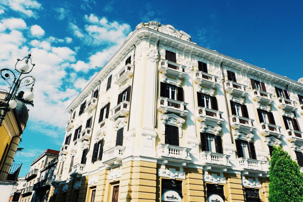 Things to see in Benevento, Campania. Corner of a large building with 4 floors, top three floors are yellow and white. Windows with balconies and wooden shutters