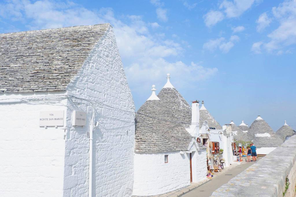 A row of trulli in Alberobello, along the left hand side are 6 trulli and along the right hand side is a wall. Some tourist are at the back of the path looking at souvenirs in a shop.