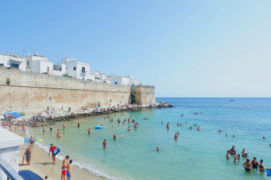 The beach in Monopoli, Puglia. The sky is blue, the sea is a deep blue / green. A tall sandstone wall runs from the left to centre of the image. Tourists are in the water swimming and on the small area of sand.
