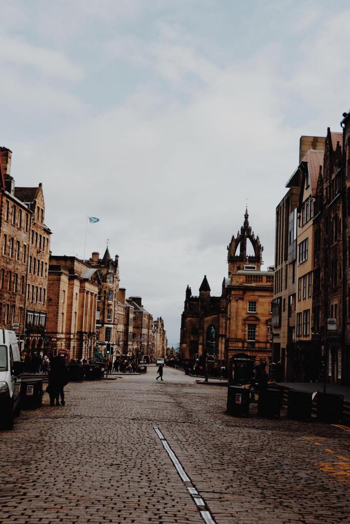 View of the Royal Mile from Castle hill. Brown buildings are either side of a cobbled road. A small Scottish flag on the left. On the right, the spire of St Giles Cathedral.