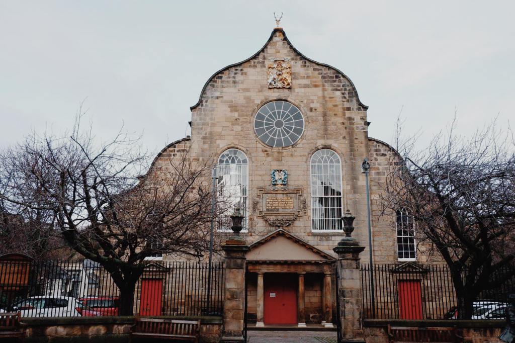 The Canongate Kirk on the Royal Mile Edinburgh. The building is tall with curved sides. At either side are brown trees in the winter. The is a red double door in the centre and two red doors either side. There are two tall windows and above one circular window.