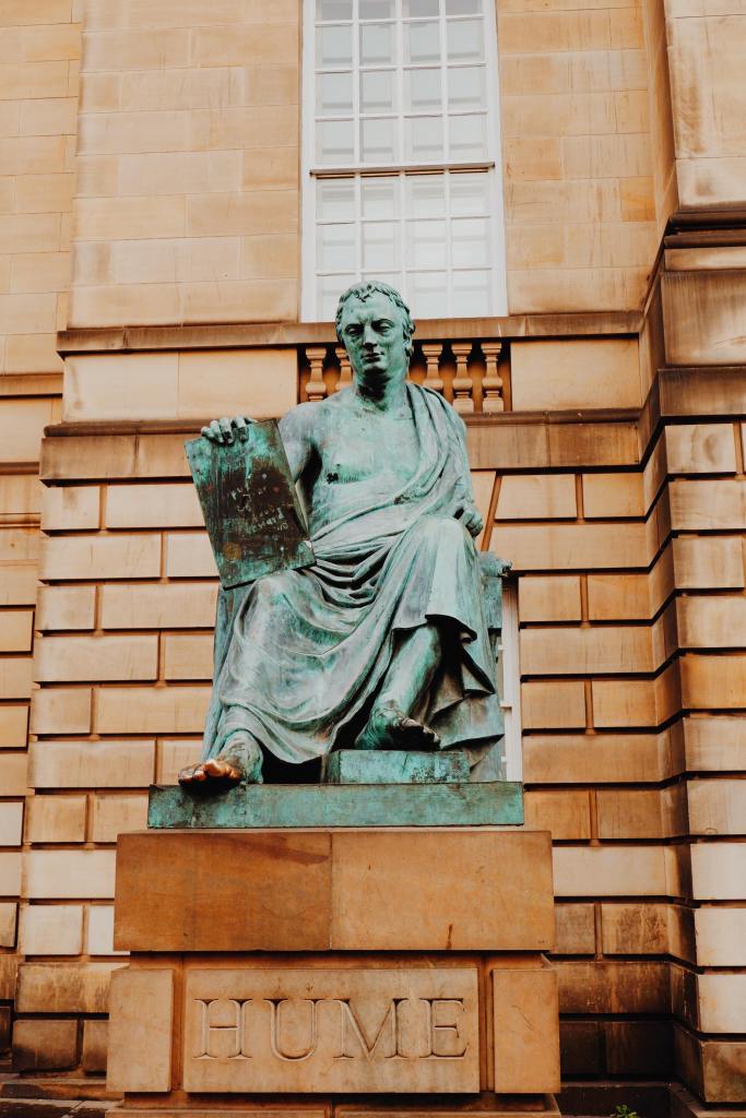 Statue of David Hume on the Royal Mile. The green statue of Hume sitting in robes holding a sign. His toe is gold. The statue sits on a carved stone saying HUME.
