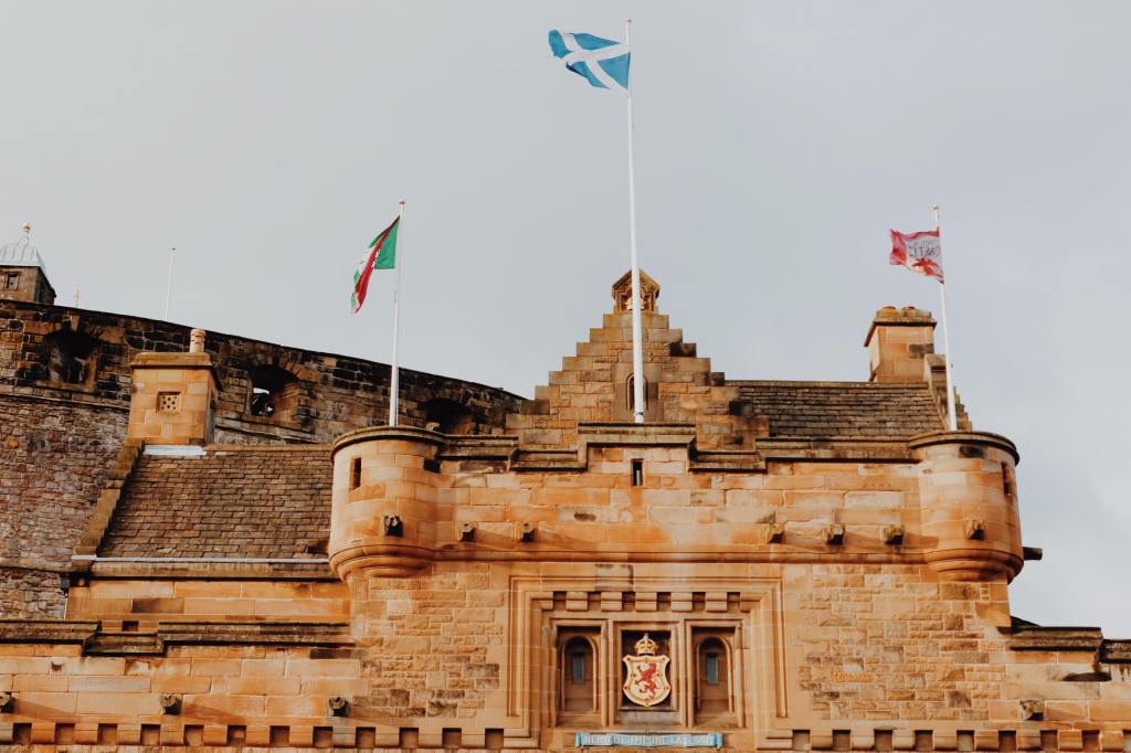 The Entrance to Edinburgh Castle with turrets. Three flags can be seen about the coat of arms. The Scottish flag in the middle. Edinburgh castle flag on the right and a green flag on left.