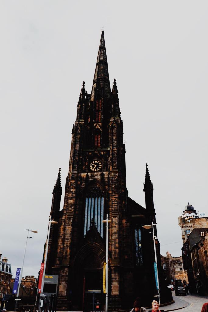 The gothic church, The Hub on The Royal Mile. The church is in the centre of the image and spans the entire length.  A clock and windows are centre of the Church. 