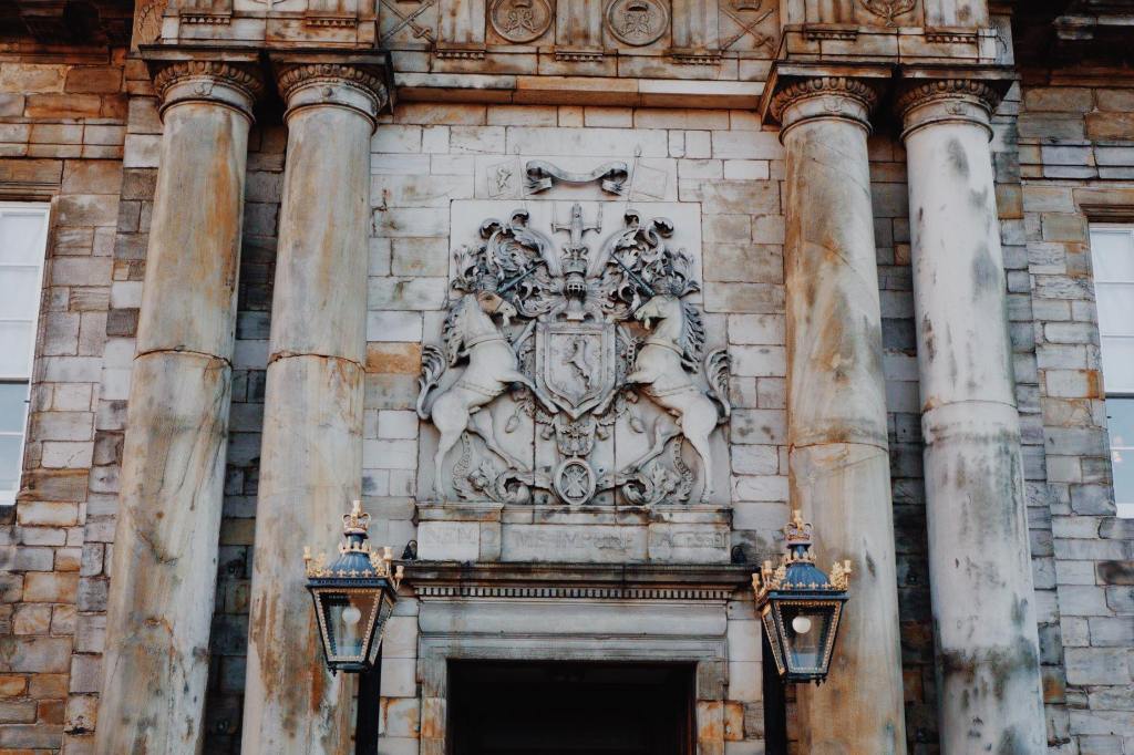 The Entrance to Holyrood Palace with the coat of arms above a door way. Two columns are either side of the door and two lampposts hang from the door.