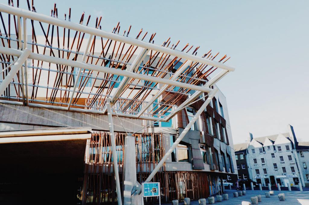 The Scottish Parliament building on the Royal Mile. The Building is white with brown wooden posts around the windows and an awning of wooden and white posts. 
