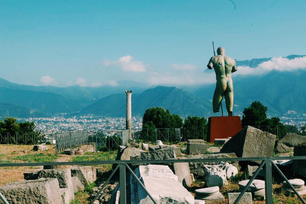 The exit of Pompeii with ruined blocks and a column. At the right is the back side of a nude bronze statue.