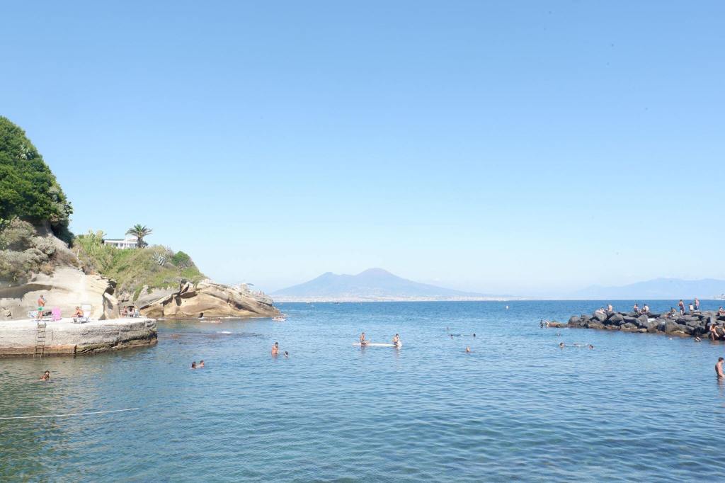 The beach at Pausilypon, Isola di Gaiola, Naples. The sea covers the lower half of the image with some cliffs and stone pier at either edge. At the horizon is Vesuvius