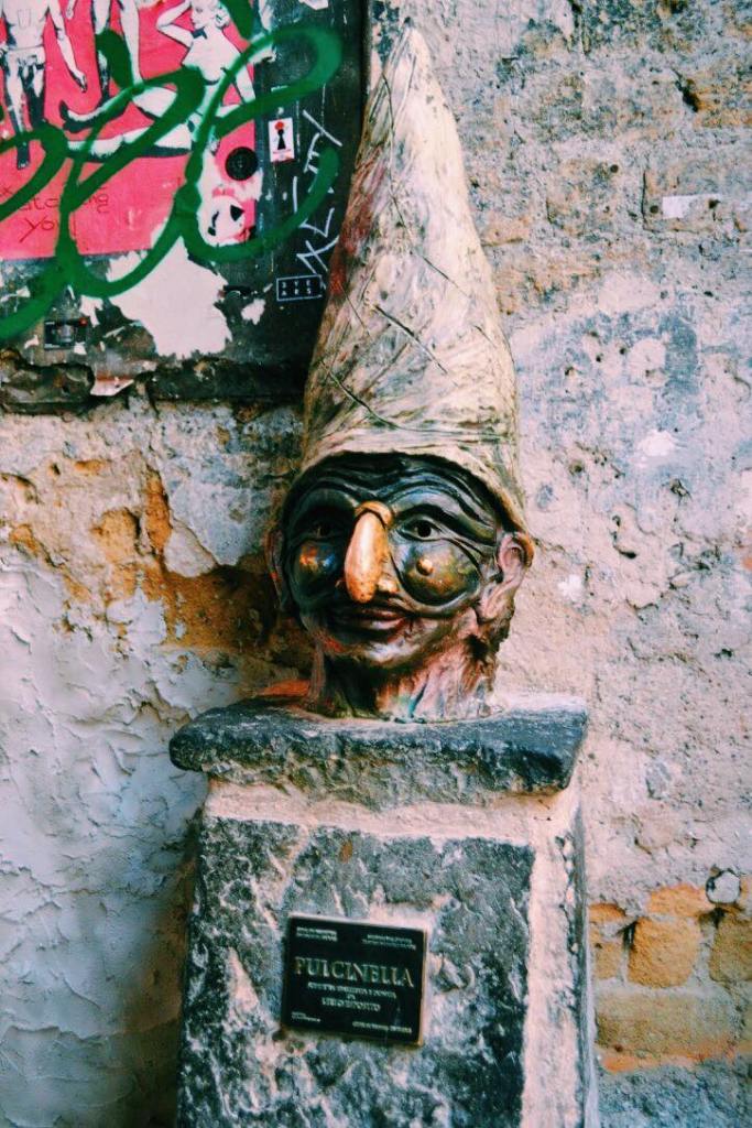 Bust of Puicinella in Naples. The statue head is on a pillar against a wall. Puicinella has a tall hat on and a black mask with a large nose. 