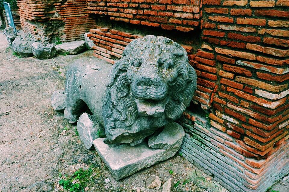Lion statue at the Roman Theatre at Benevento. The lion has no front legs and is crumbling. It rests against a red brick wall.