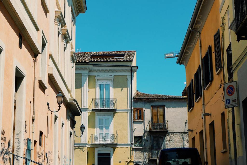 Street in Benevento Campania. Buildings. Either side of the street are yellow and orange buildings and at the end of the road are white and yellow houses with balconies. 