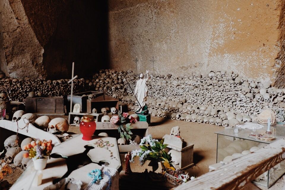 Skulls in rows on rows along the tufa walls, with rows of long bones onto. Statues of Jesus, crosses and flowers are scattered amongst the remains. A coffin is at the front of the image with beads on top.