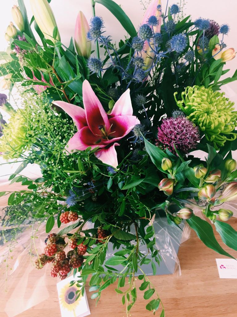 A large bunch of flowers with a purple Lily in the middle and lots of leaves and greenery 